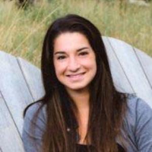 A person smiling while sitting outdoors on a wooden chair. This alt text was added with Al; accuracy may vary.