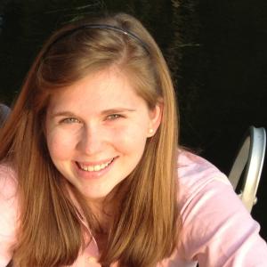 Smiling person in sunlight wearing a pink shirt, sitting near water. This alt text was added with Al; accuracy may vary.