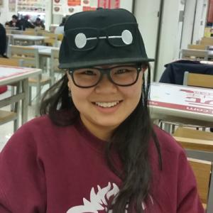 Smiling person wearing glasses and a black hat, sitting in a cafeteria. This alt text was added with Al; accuracy may vary.