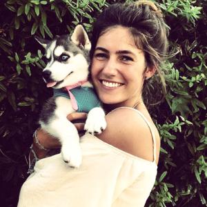 Person smiling while holding a small dog, surrounded by greenery. This alt text was added with Al; accuracy may vary.