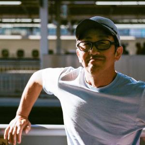 Man in grey shirt and dark hat with glasses standing outside in the sun