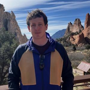 Man with short brown hair and a jacket in front of mountains and a blue sky