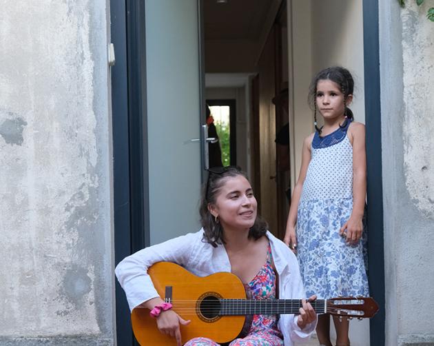 A student sits outside on stone stairs playing the guitar while a child watches from the top of the stairs. 