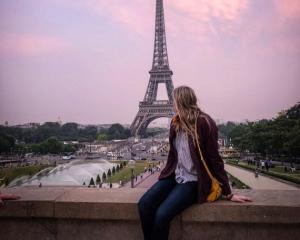 a student girl sitting on a bridge and looking at the eiffel tower with a pink sky on the background 
