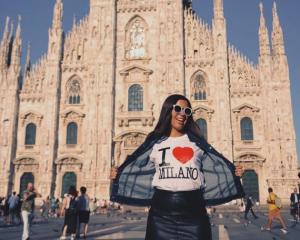 a girl student standing in front of a historical site building and showing off her shirt with words " I love Milano"