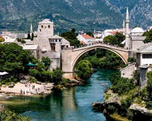 A bridge connecting two sides of a Mostar arches over a slow-moving, shallow body of water. In the background are verdant mountains.