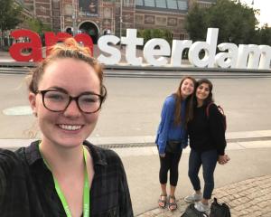 three students pose in front of a white and red sign that reads "amsterdam"