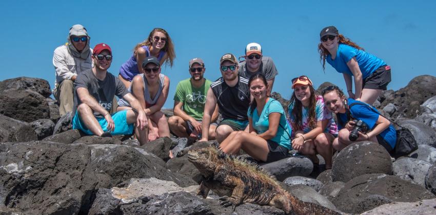 a group of students posing for a photo atop volcanic rock with a marine iguana