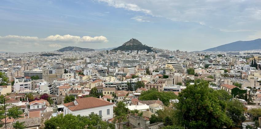 Distant view of a cityscape (Athens) with scattered green trees and a mountain under a cloudy sky.