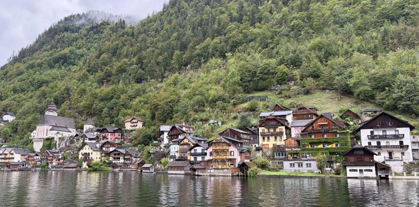 Some small colorful houses on Lake Hallstatt. The hill behind them is covered in green trees, with fog in the distance.