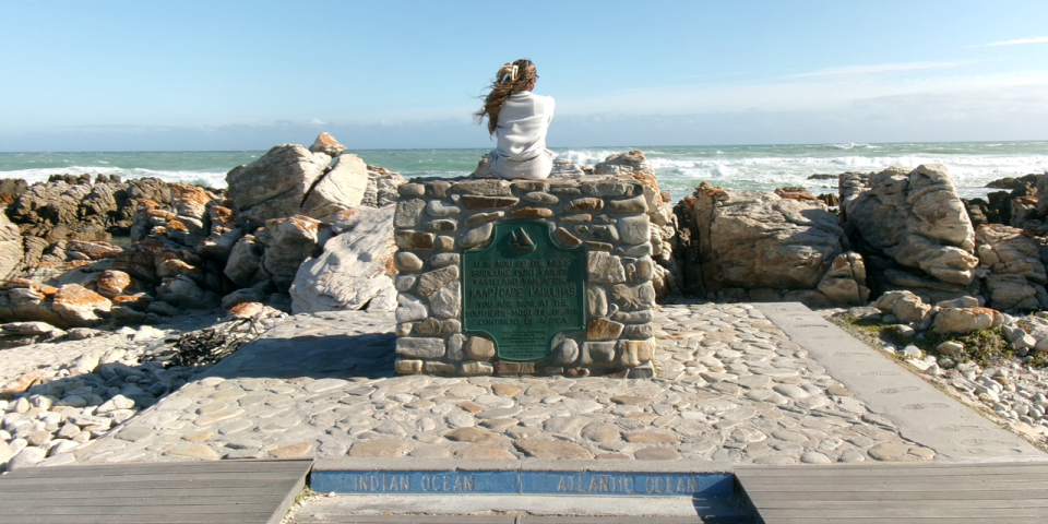 Picture of me sitting on a structured pile of rocks with a plaque reading that where I am is Africa’s southernmost point. East of this is the Indian Ocean, and west is the Atlantic Ocean. 