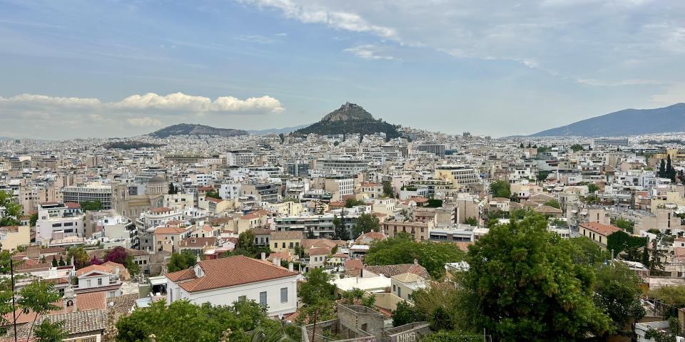 Distant view of a cityscape with scattered green trees and a mountain under a cloudy sky. This alt text was added with Al; accuracy may vary.